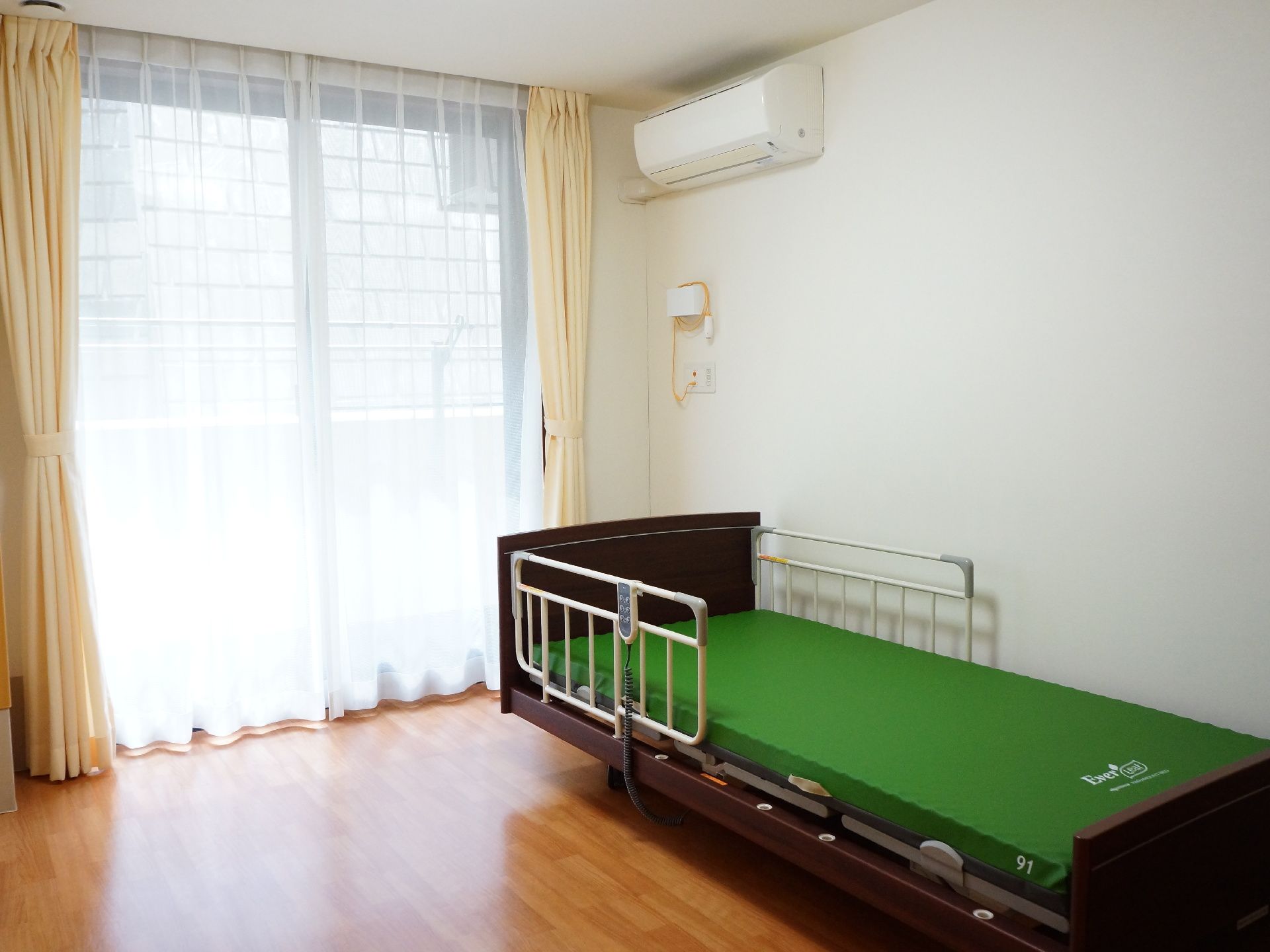 窓にはカーテンがかかり光が差し込んでいる。右上にはエアコンが敷設され壁に寄せて可動式のベッドが置かれている。