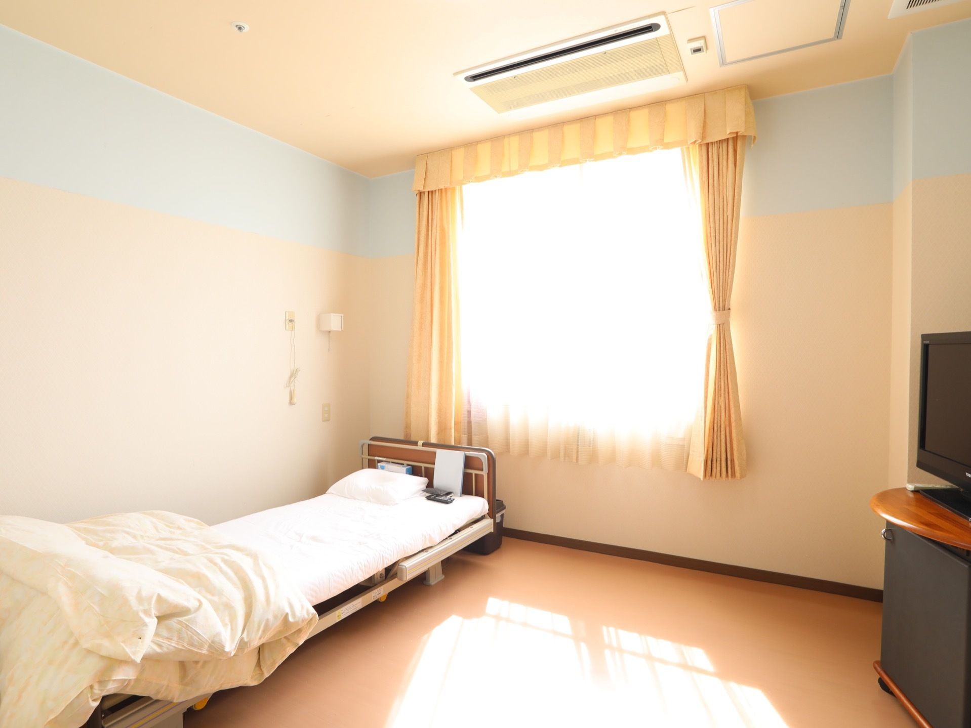 レースのカーテンを通して日光が差し込む明るい部屋である。壁際にシーツをセットしたベッドがある。エアコンが天井に設置されている