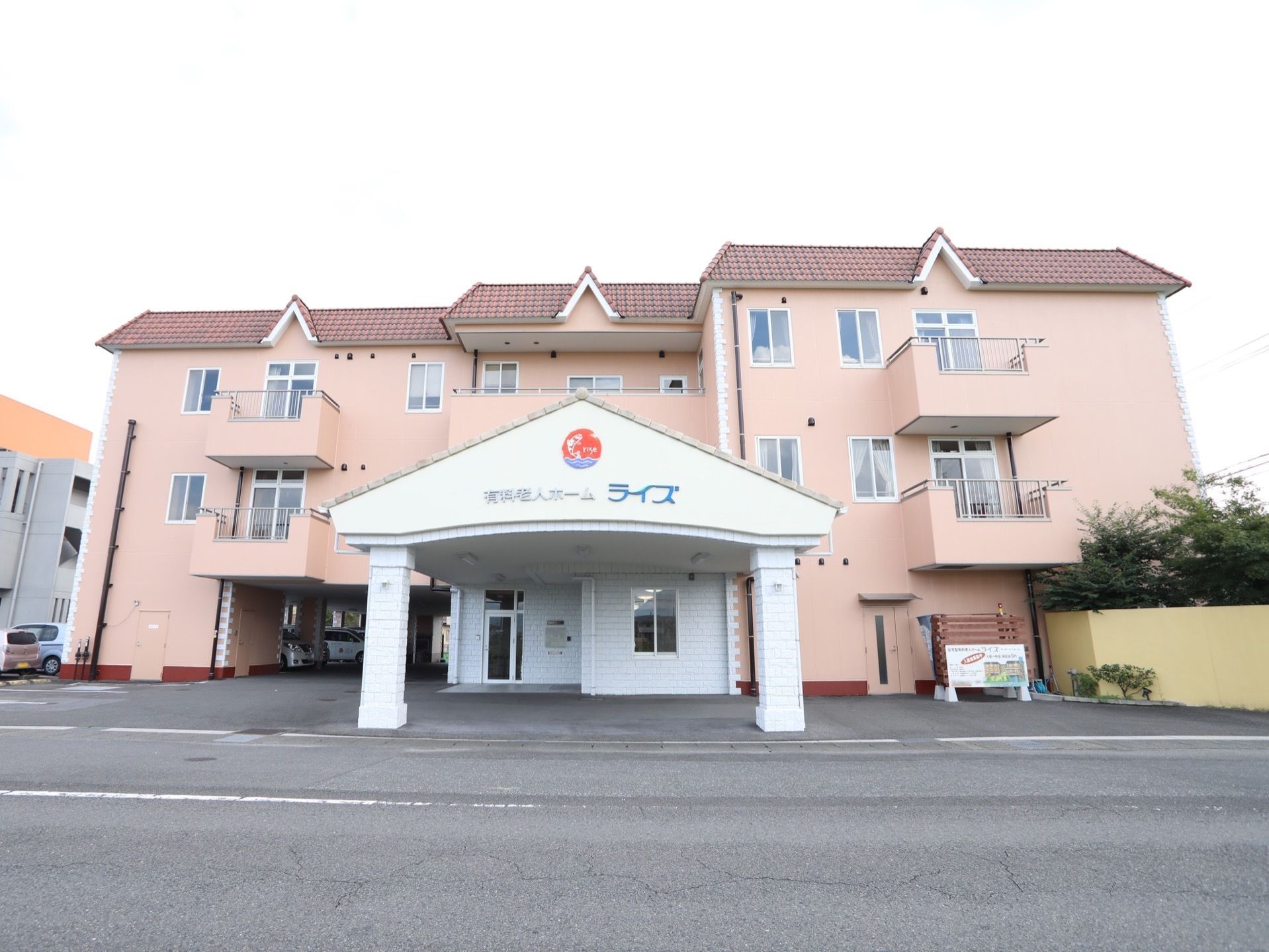 施設を正面の公道から見た様子。中央に三角屋根の玄関エントランスがある。建物は3階建てで、１階左端は通り抜けの車庫の構造である。