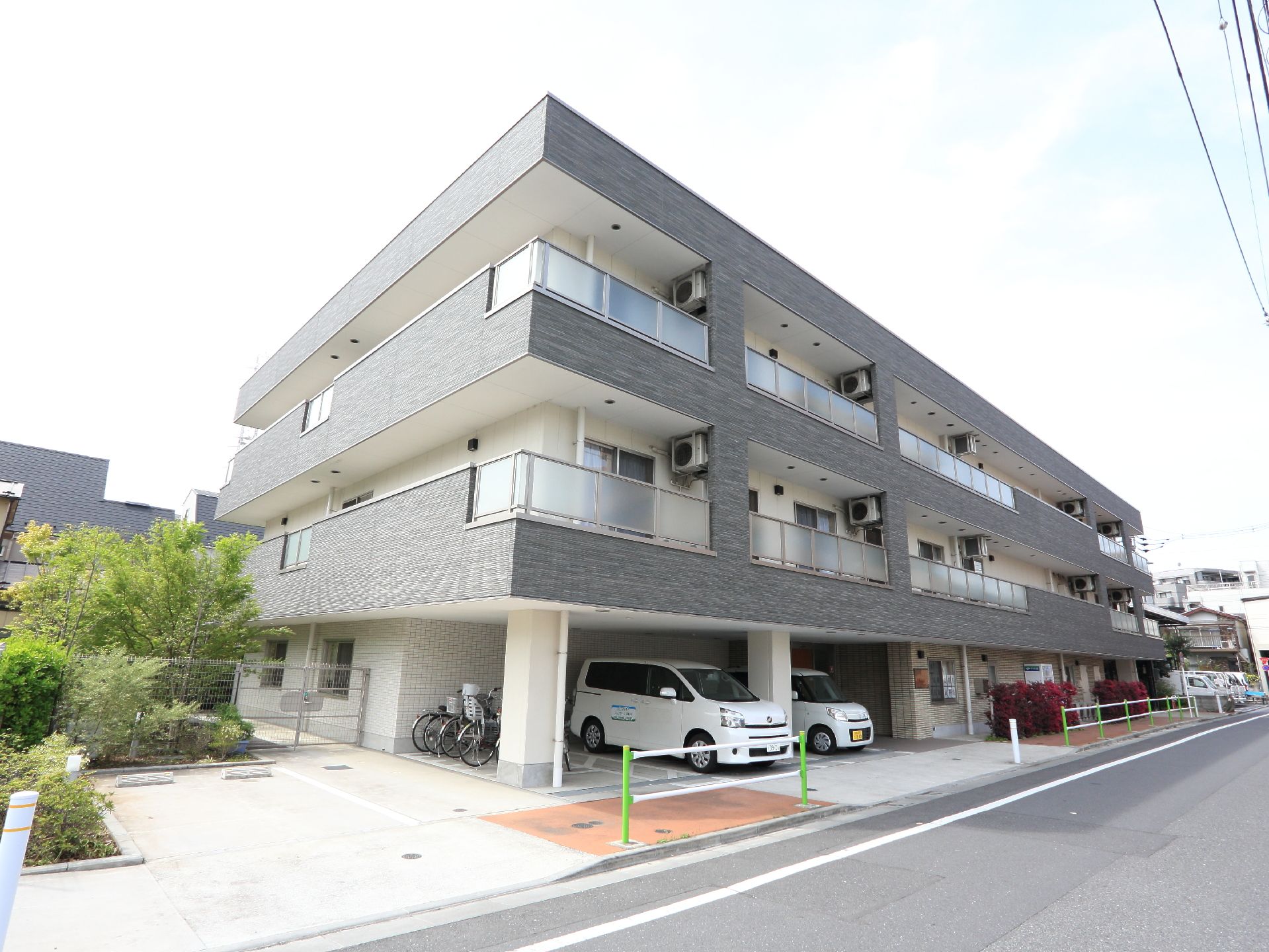 グレーと白で統一された3階建ての建物。道路に面している。施設の横には木が植えられている。1台分の駐車スペースが確保されている。