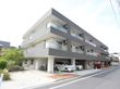 グレーと白で統一された3階建ての建物。道路に面している。施設の横には木が植えられている。1台分の駐車スペースが確保されている。