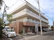 ブラウン系の色をした3階建ての施設。マンションのような外観。施設の看板が道路沿いに設置されている。複数の駐車スペースがある。