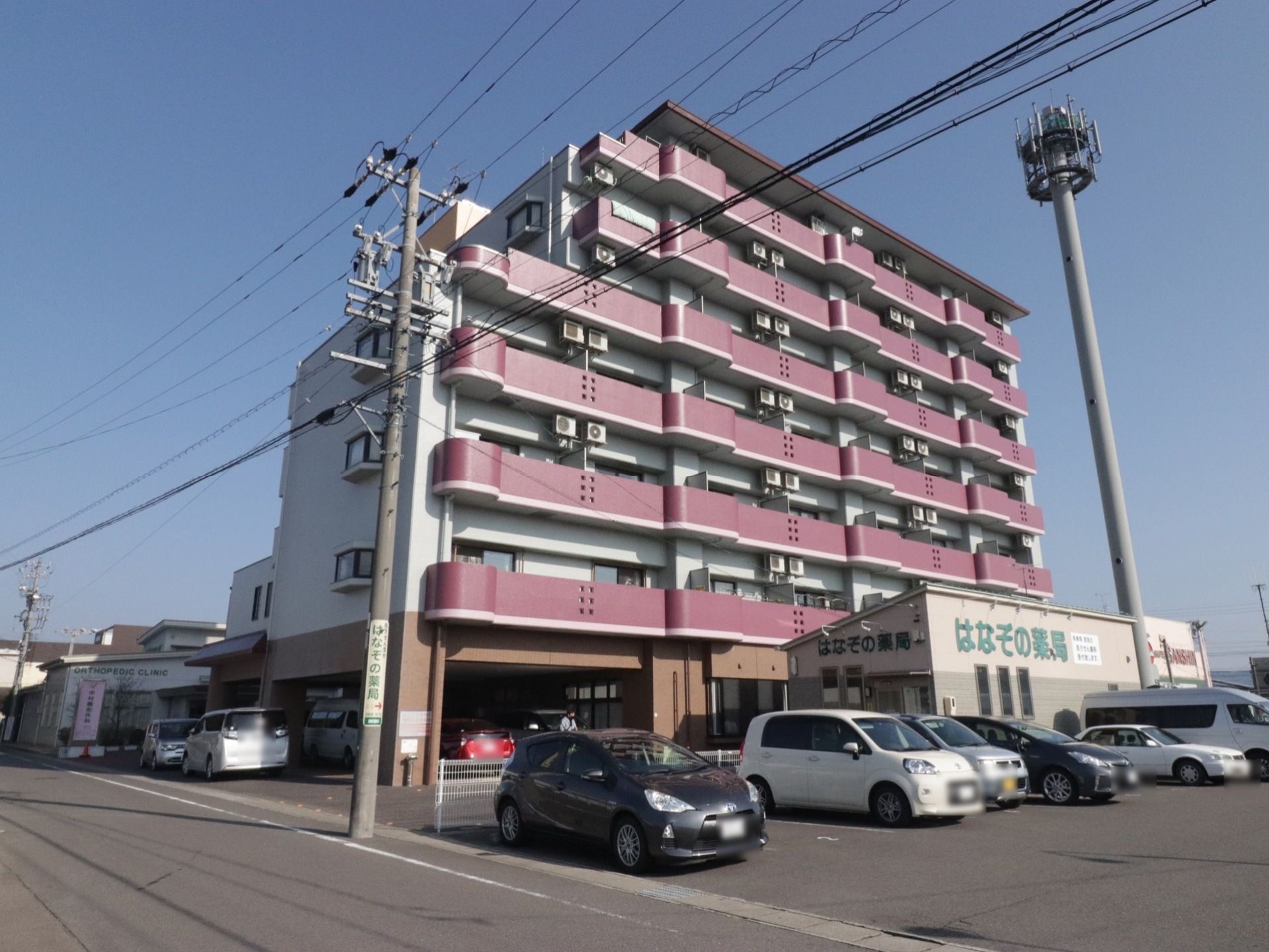 白色を基調に、エンジ色のフェンスが突き出た７階建ての外観である。薬局が隣接し、駐車スペースも完備している。