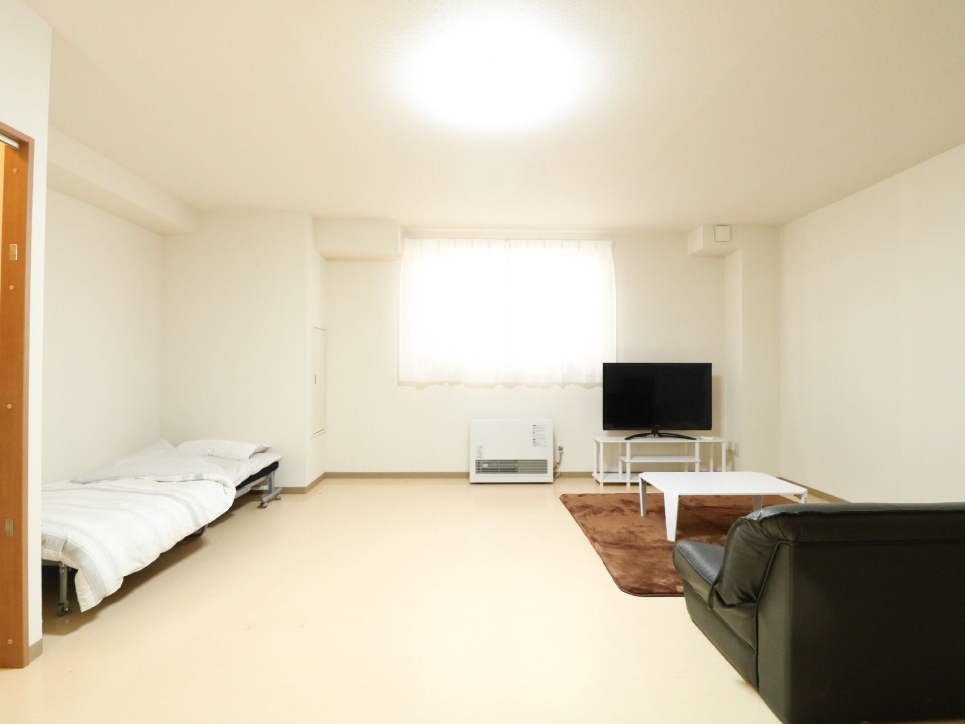 白色を基調とした広々とした室内。左側にベッドがあり、右側にテレビやラグ、テーブルとソファが配置されている。