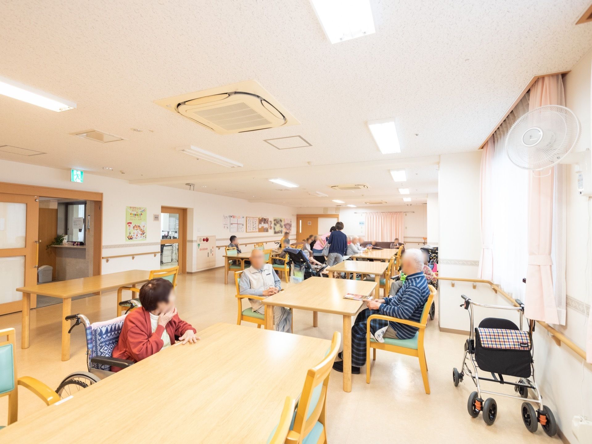 広い食堂に、テーブルがゆったりと配置されている。車椅子をご利用の女性の入居者様が手前のテーブルについている。