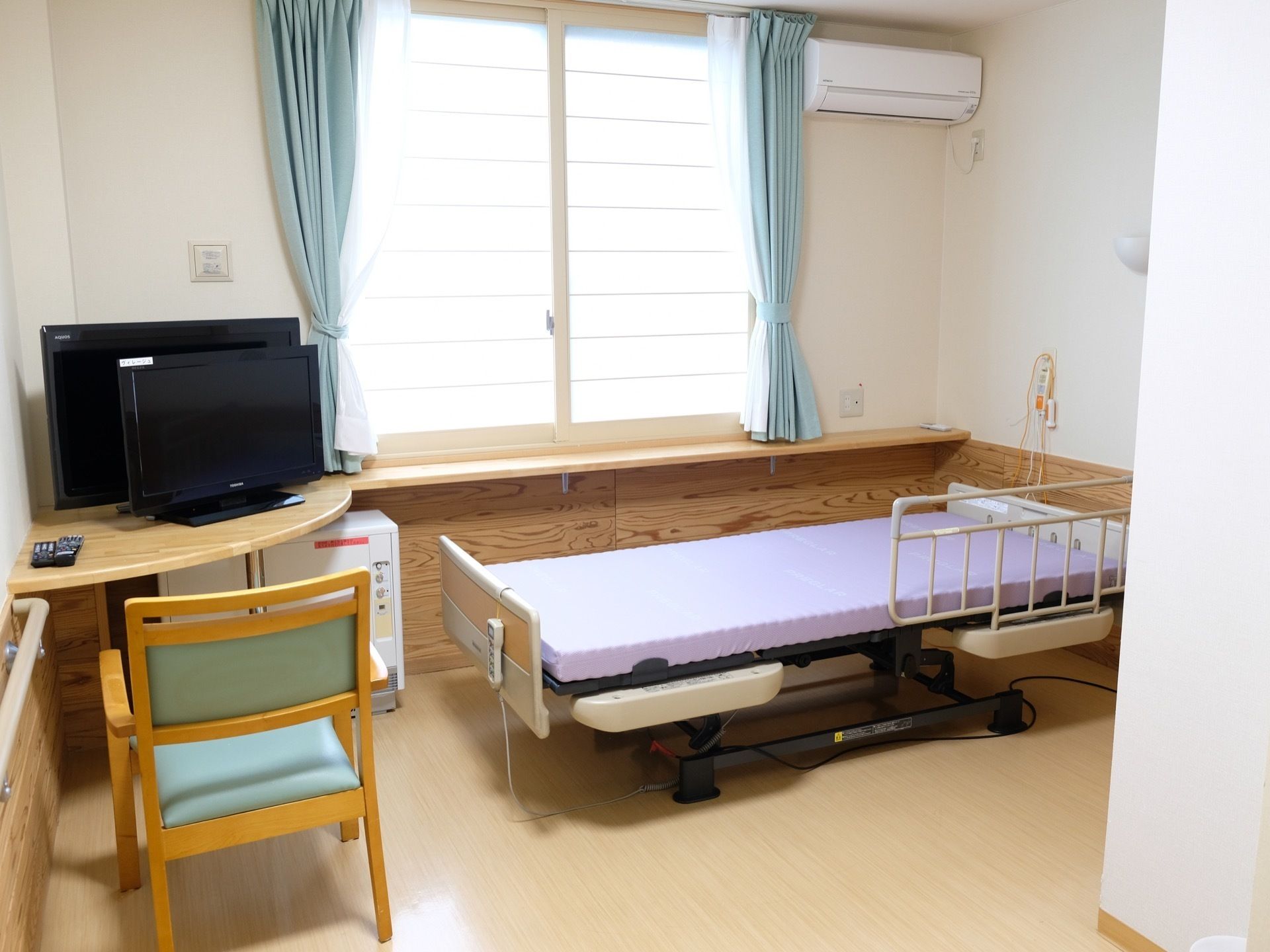 窓際に木製の台が設置されていて、その横にベッドが置かれている。手前には部屋を仕切るカーテンが吊られている。