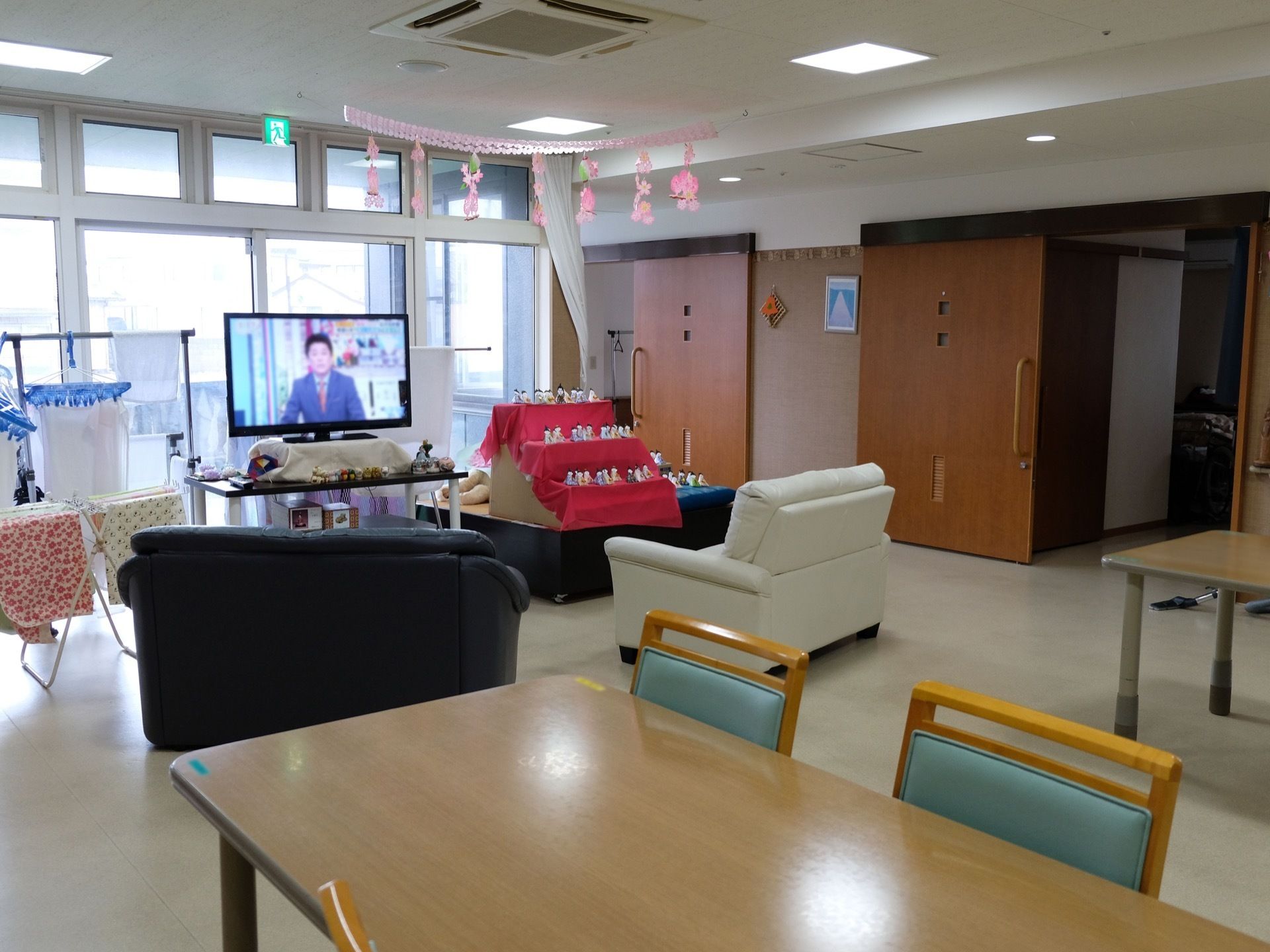 窓際に雛祭りの飾りがある。その手前には白とグレーのソファが置かれている。大型のテレビが設置され、その横に洗濯物が干してある。