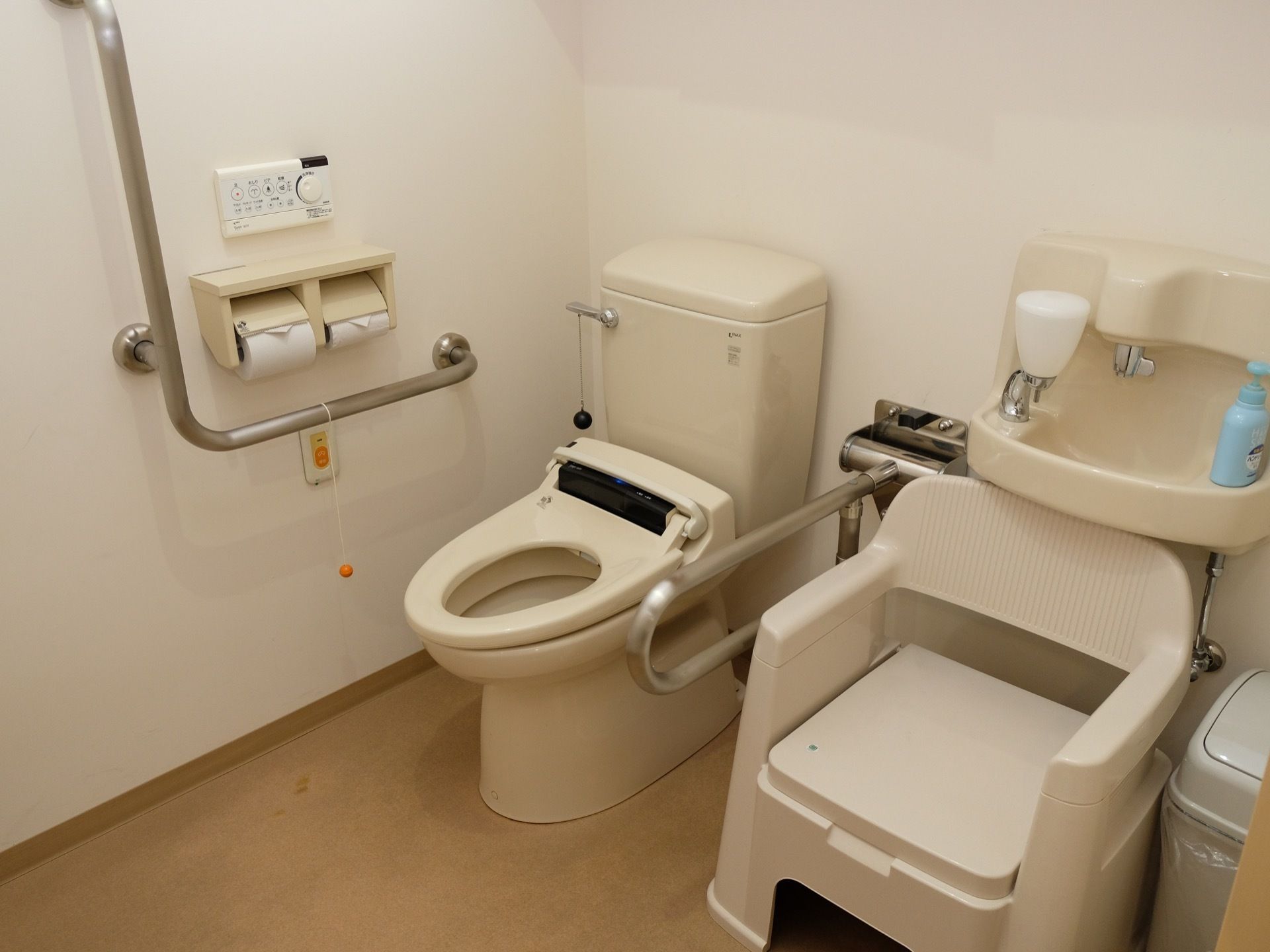 ベージュ調の床と白い壁に囲まれたトイレである。アイボリーの温水洗浄便座が設置され、簡易トイレも置かれている。