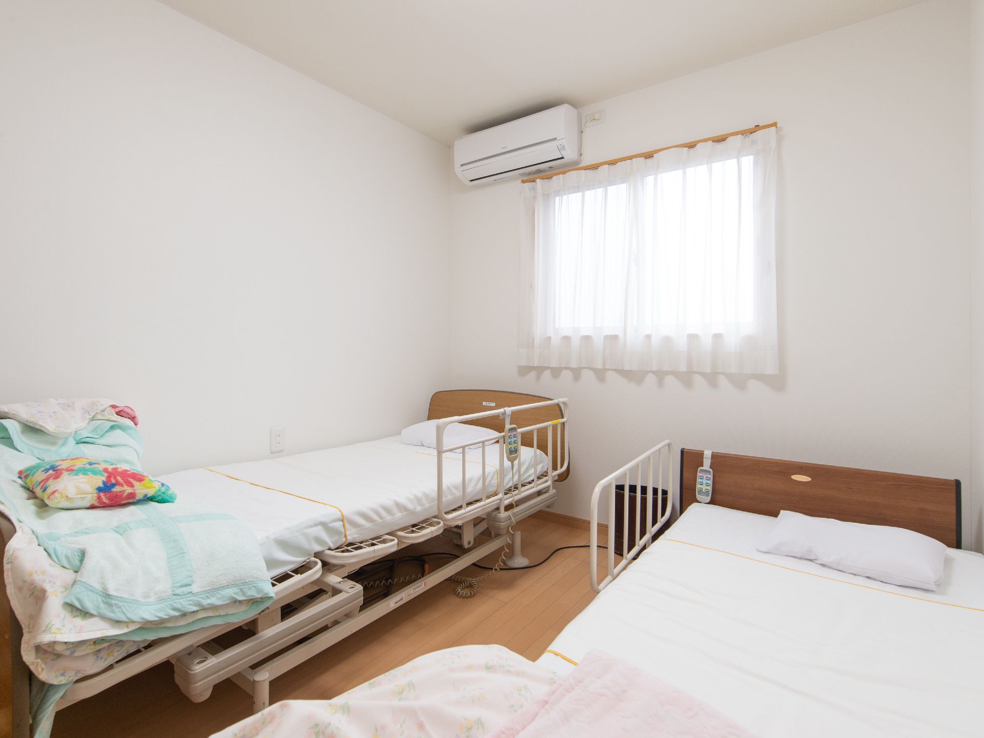 白い壁と明るいフローリングの部屋に、介護用ベッドが２台並べて設置されている。窓があり、エアコンを完備している。