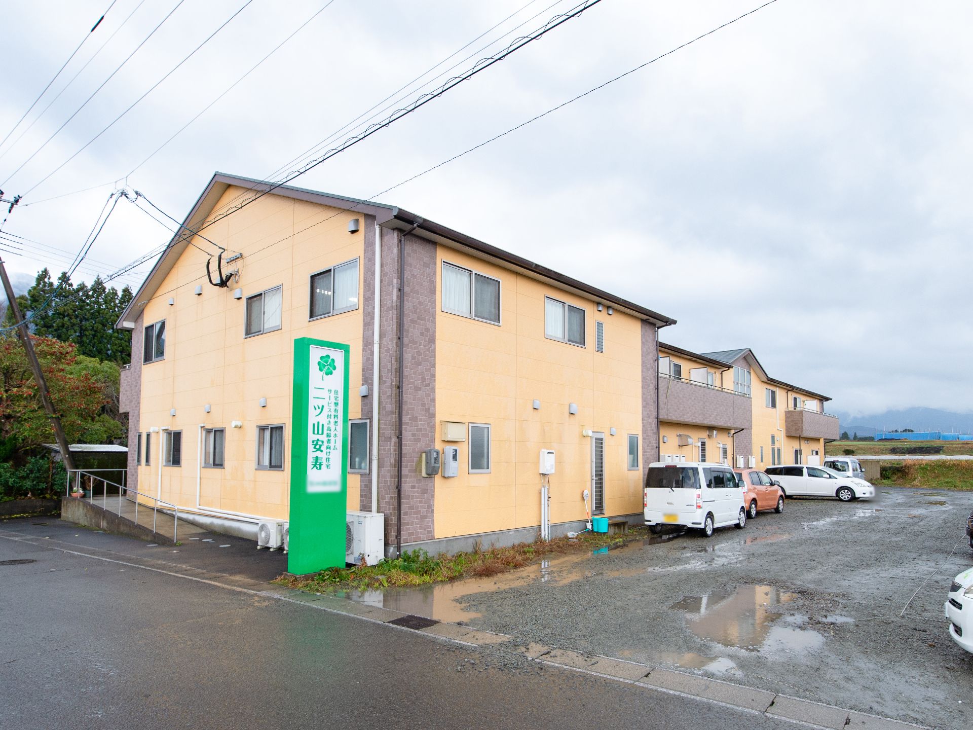 黄色い壁の可愛らしい２階建ての建物である。建物の隣には広く駐車スペースが設けられており、自動車が数台停まっている。