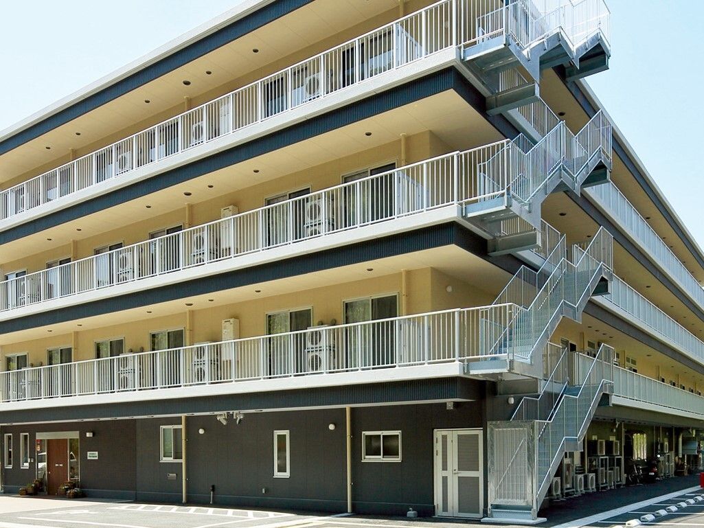 ダークブラウンとオフホワイトのマンション風の建物である。施設に隣接して広い駐車スペースが設けられている。