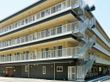 ダークブラウンとオフホワイトのマンション風の建物である。施設に隣接して広い駐車スペースが設けられている。