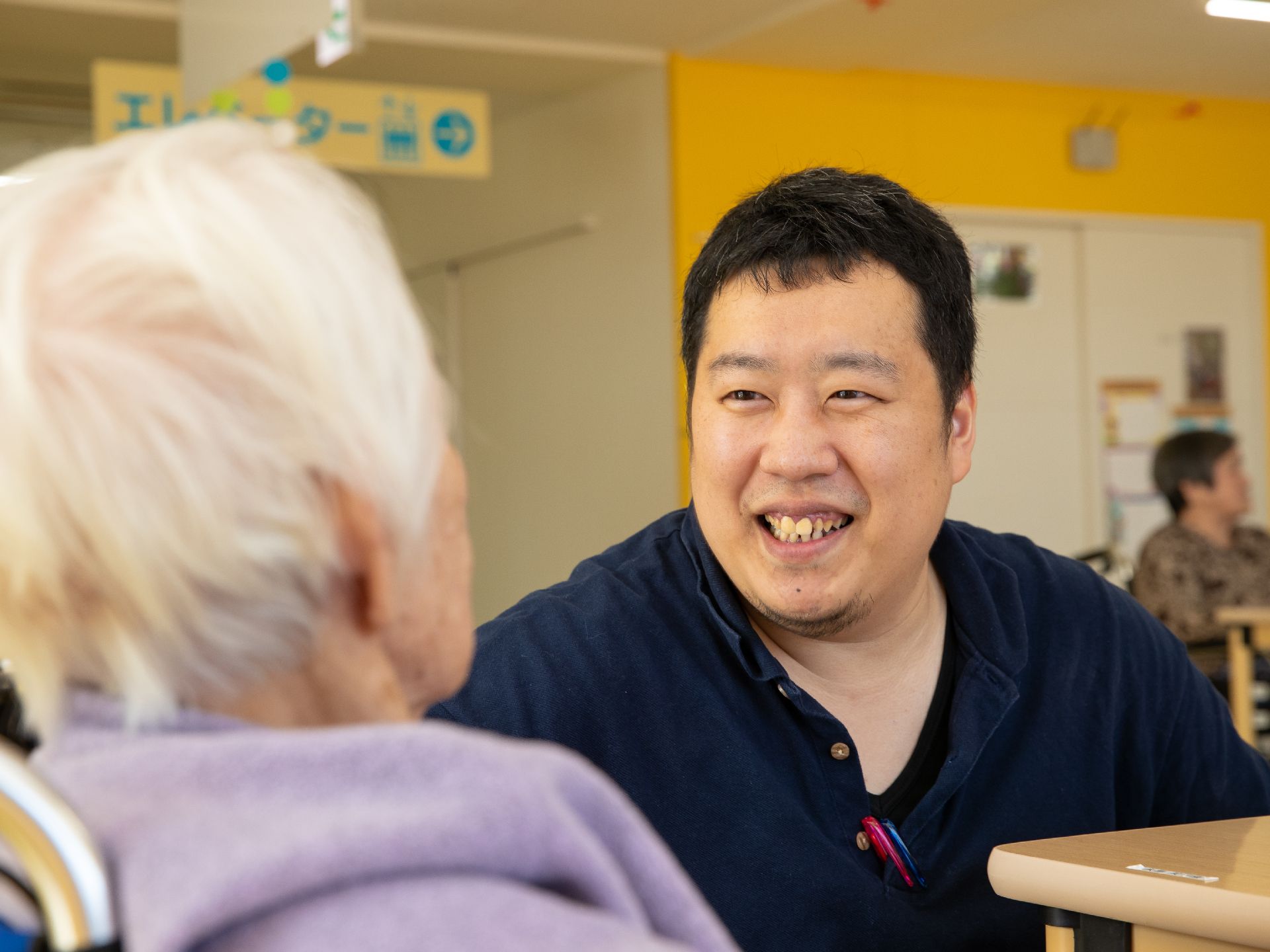 男性スタッフが笑顔で入居者様と会話をしている。目線を合わせてにこやかな表情である。一人一人に合ったサポートを行っている。