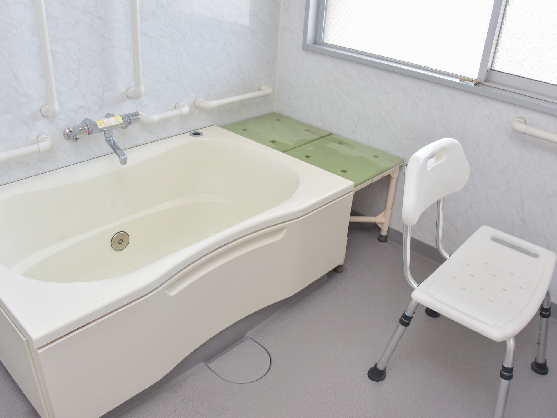 薄いグレーの床、白い浴槽。白い介護用の風呂椅子が設置。壁には手すりがある。大きい窓からは光が入る設計。