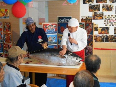 大漁旗をイメージさせる衝立を背に、台に乗った大きなマグロの解体が始まろうとしている。入居者様は本格的なショーを間近で観賞する事ができる。