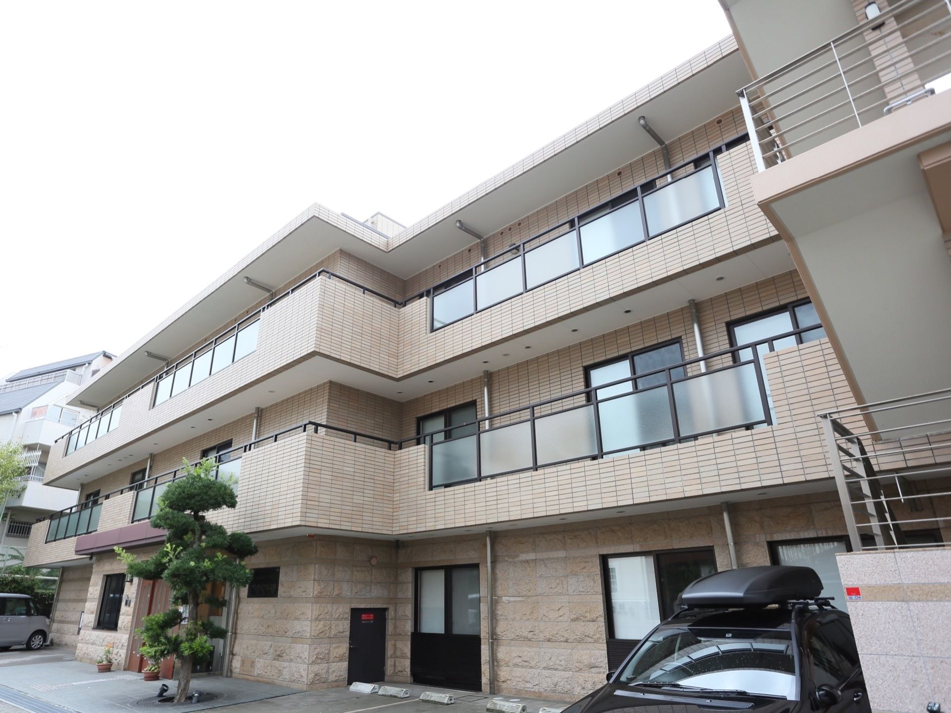 ベージュを基調とした３階建ての施設である。一番上は屋上になっている。建物の前方に駐車スペースを設けている。