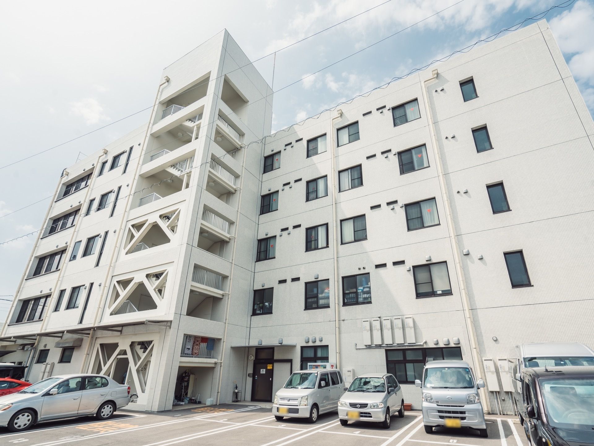 白い外壁の5階建ての建物で、外側に非常階段が設けられている。施設前の路面は舗装され、駐車場として利用されている。