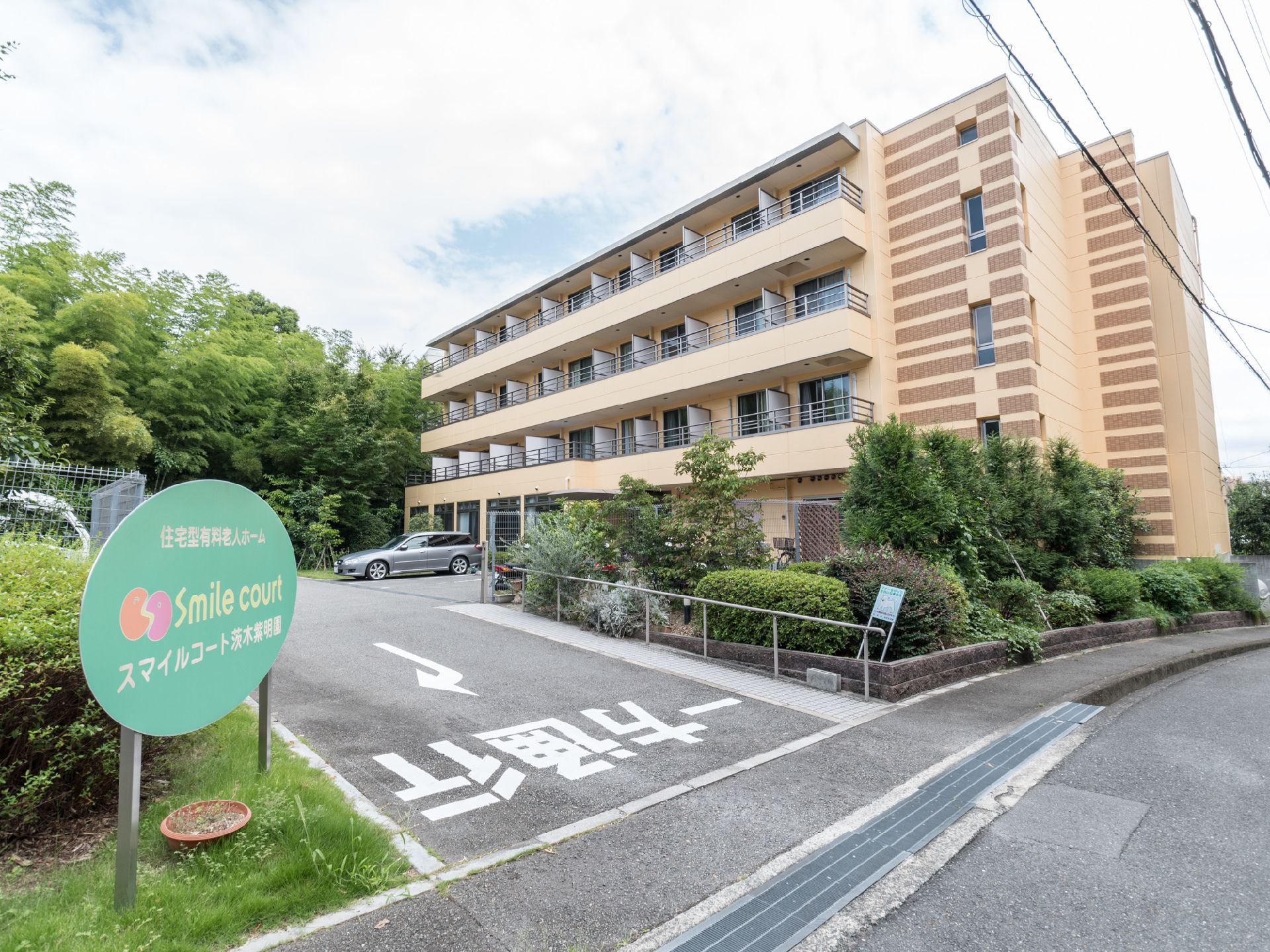 施設名を示す看板が設置されている。路面に一方通行と書かれている。建物の前に自動車が停車している。たくさんの植樹がされている。