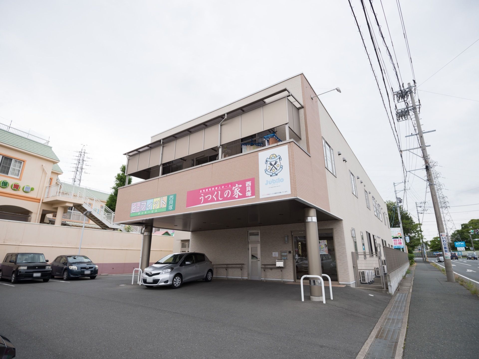 2階建てのピンク系の建物である。エントランス前には駐車スペースがあり、広い道路に面している。周辺はアスファルトで整地されている。