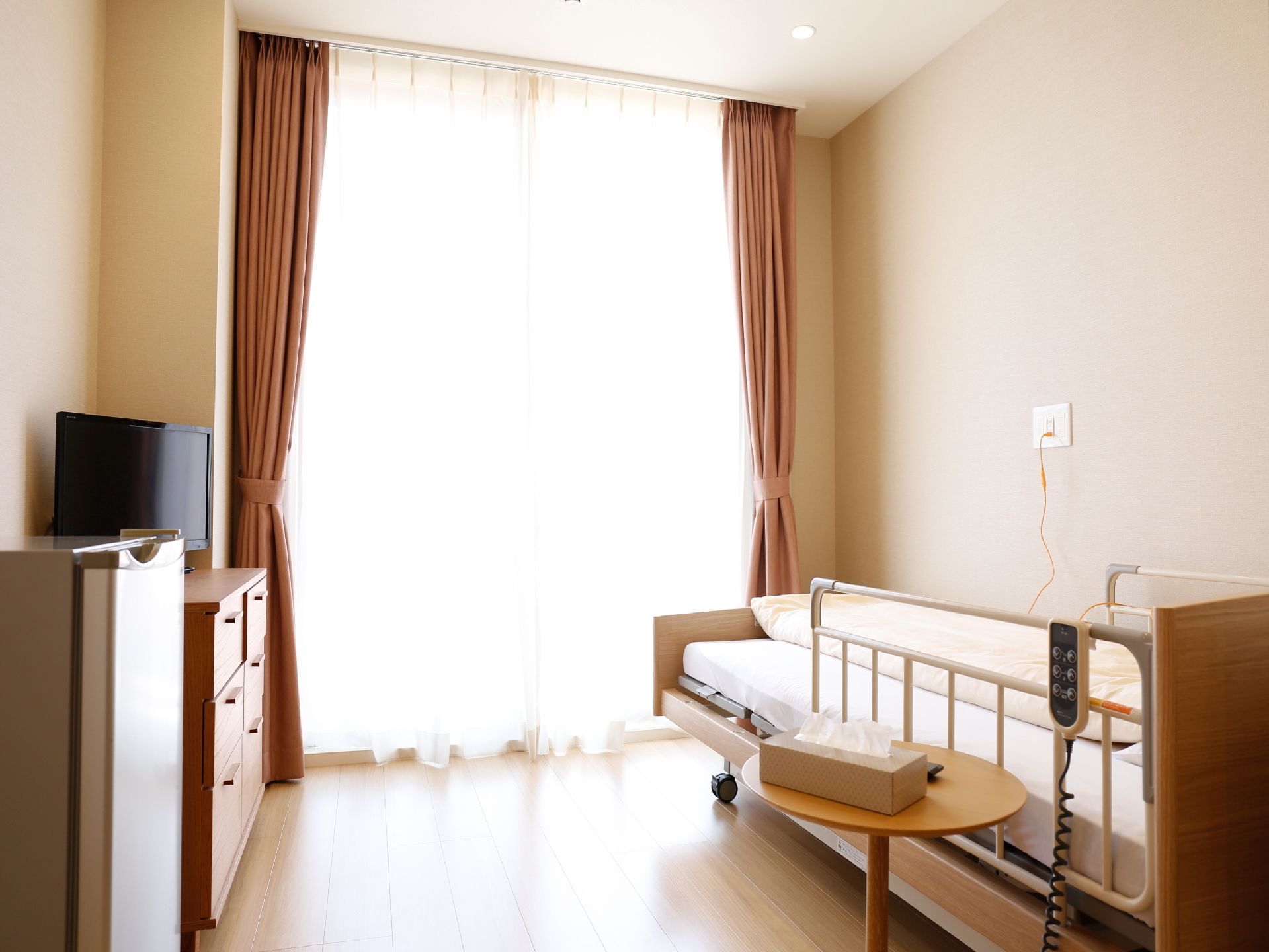 床は木目調のフローリングで天井まである掃き出し窓がある。壁に沿って低めの手すりつきベッドがある。反対側にチェストがありその上にテレビがある。