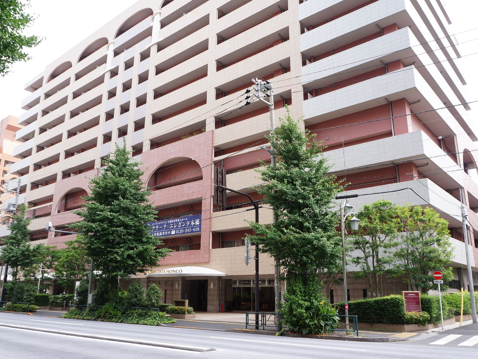 建物正面前には片側2車線の道路と歩道がある。歩道と建物の間に低木の植栽帯があり、所々に高木が配置されている。