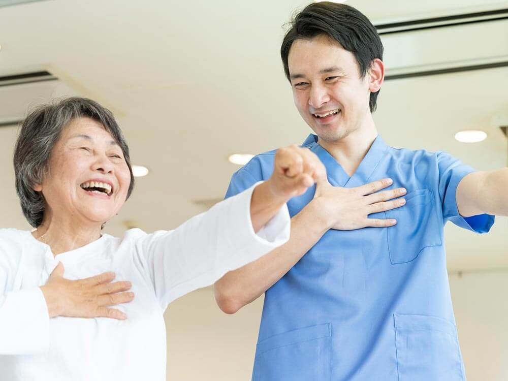 青いユニフォームを着た男性スタッフと白い服を着た高齢女性が笑顔で体操をしている。