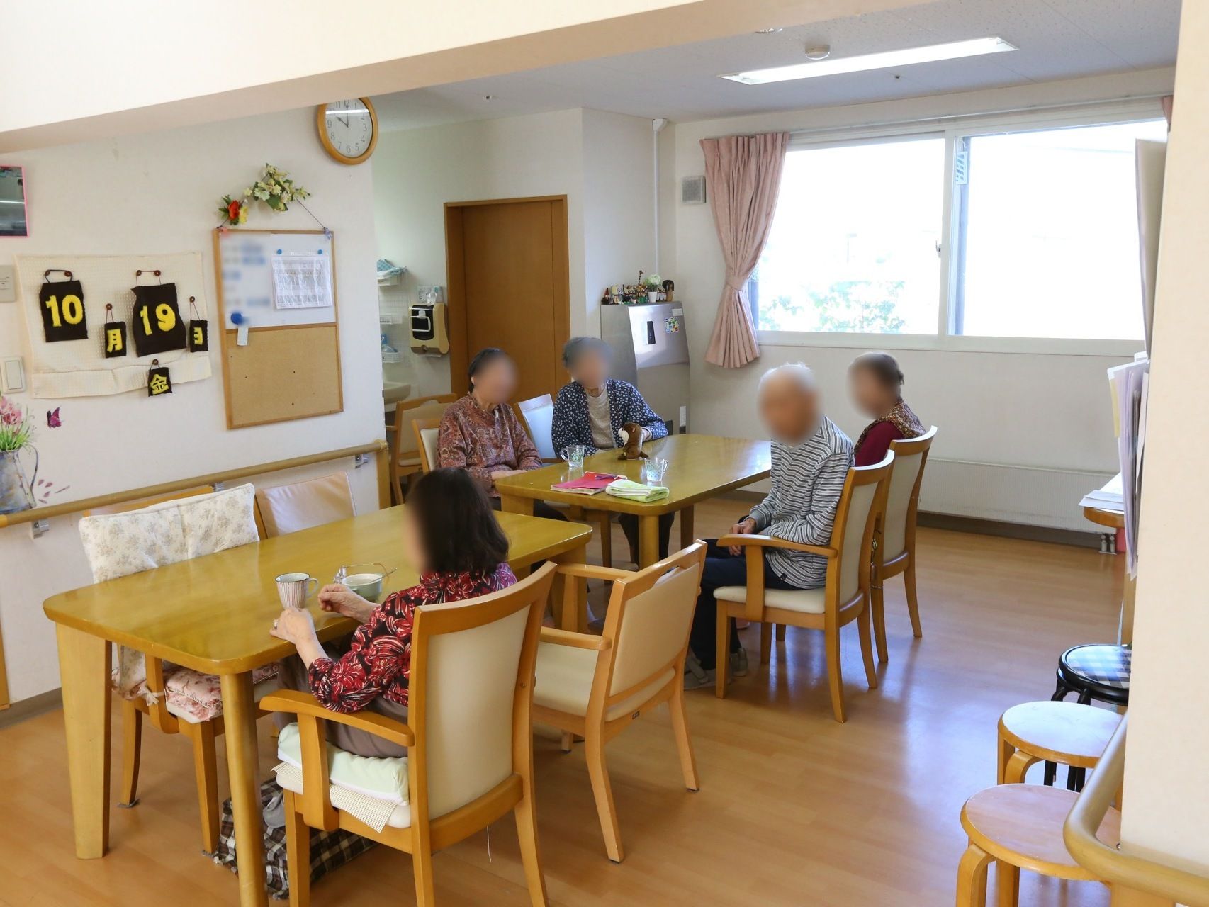 ダイニングテーブルのセットが置かれており、入居者様たちが座ってくつろいでいる。壁に手作りの日付カレンダーが飾られている。