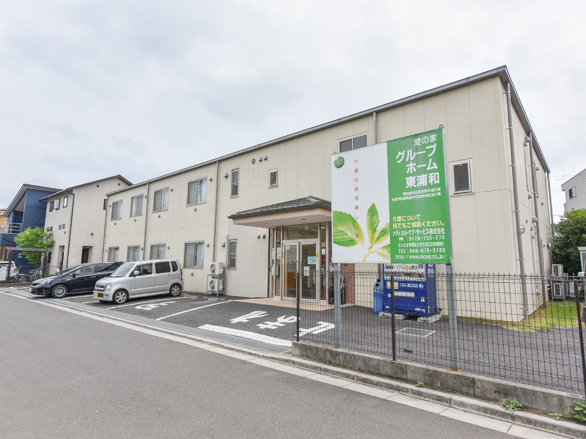 施設手前左側には駐車場あり。玄関の右側には自動販売機が置かれている。フェンスには施設の案内が書かれた看板が取り付けられている。