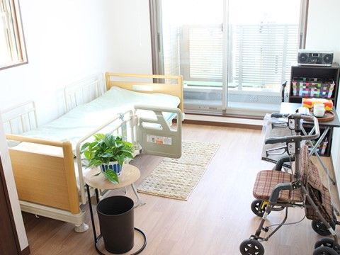 掃き出し窓があり、ベランダが設置されている。室内にはベッドやテーブル、収納ケース、歩行器などが置かれており、床はフローリングである。