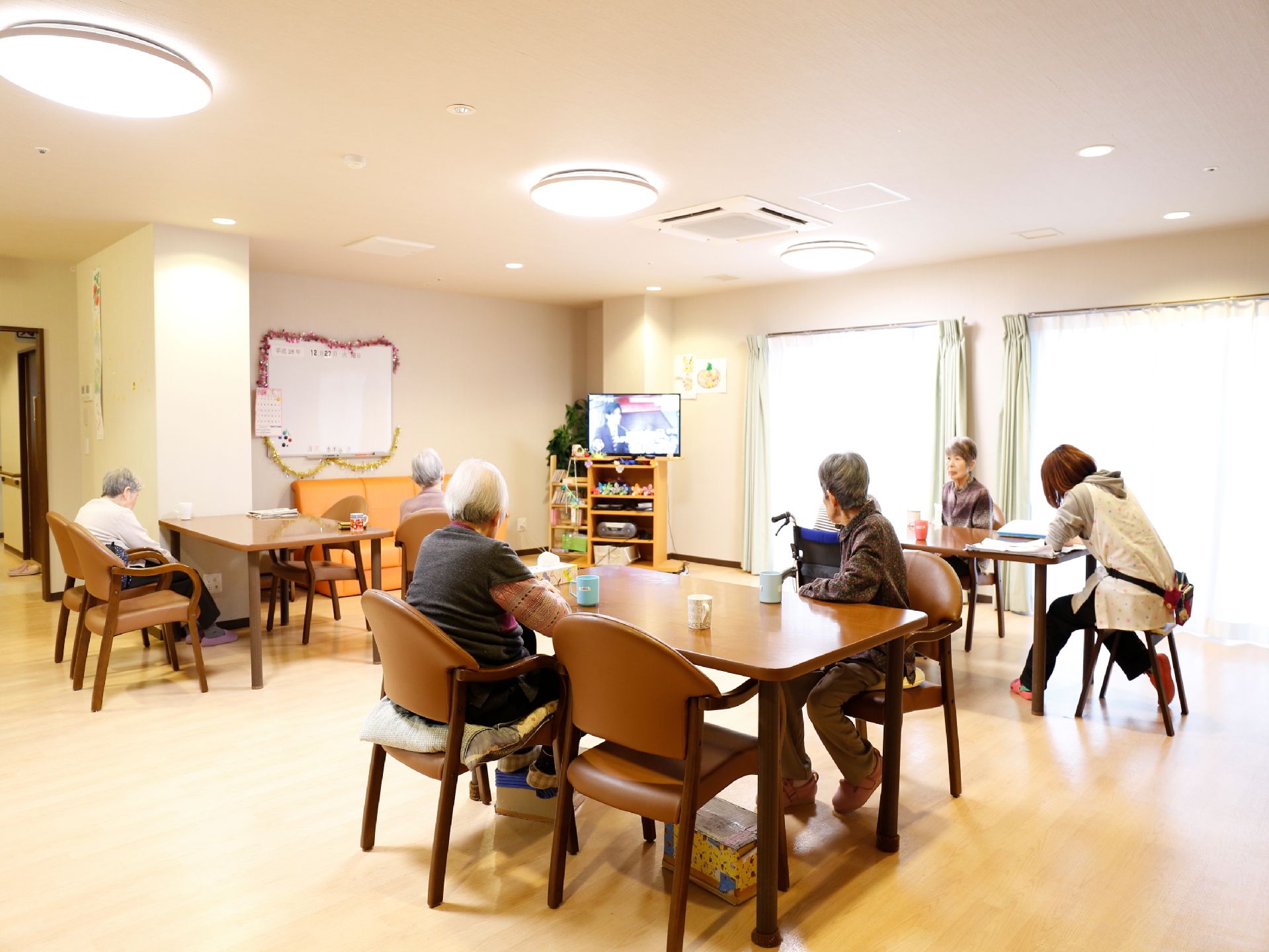 共用スペースに居住者が集まってテレビを観ている。茶色い机が3か所に分かれており、それぞれ椅子が4脚並べられている。
