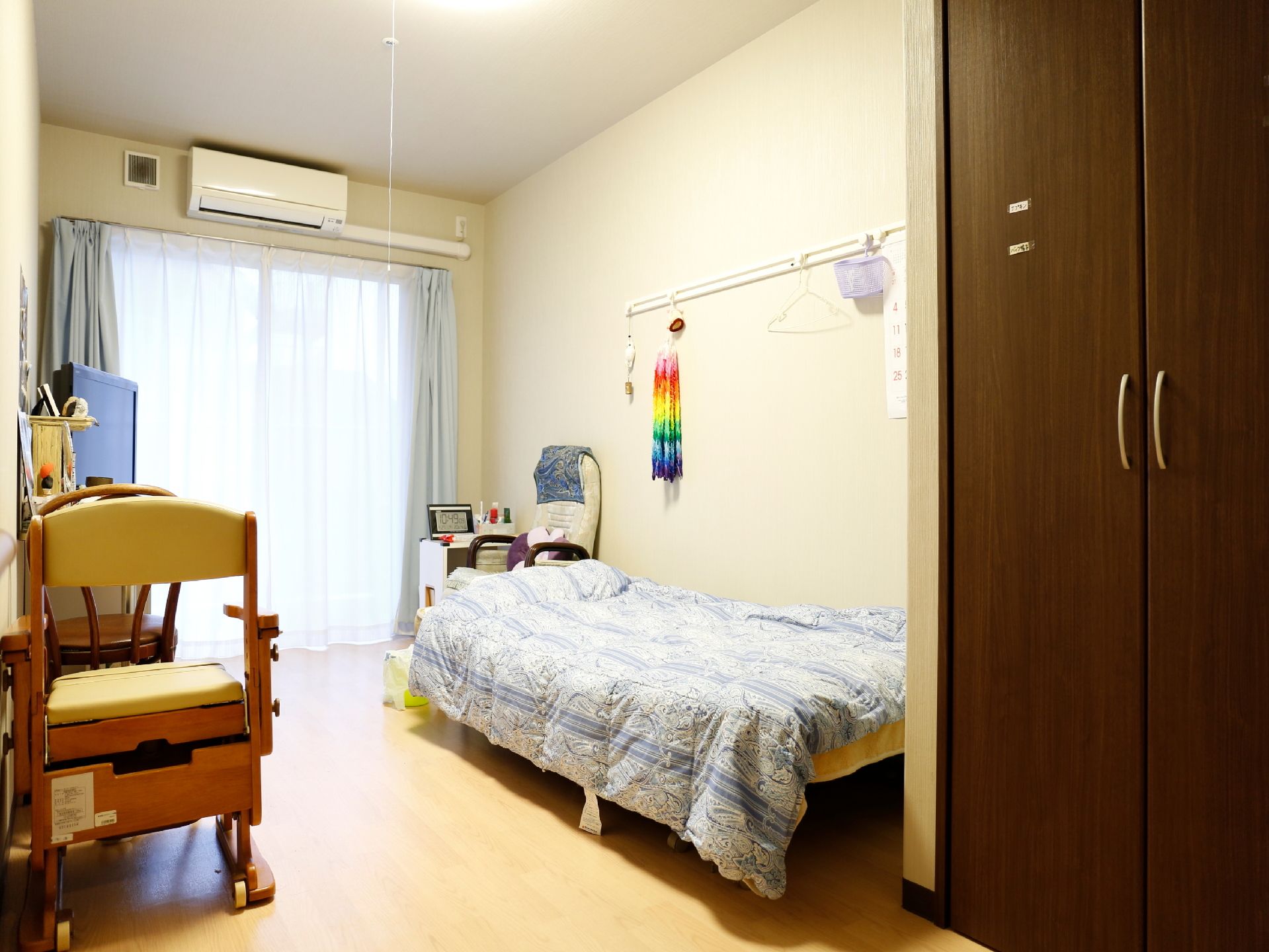 ある居住者の居室。壁にはフックが取り付けられているため、千羽鶴やカレンダーが飾ってある。ベッドの隣には座椅子が置いてある。