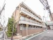 通学路に面した通りの角に施設建物が建っている。すぐそばに商店街がある。1階部分に植え込みや勝手口がある。
