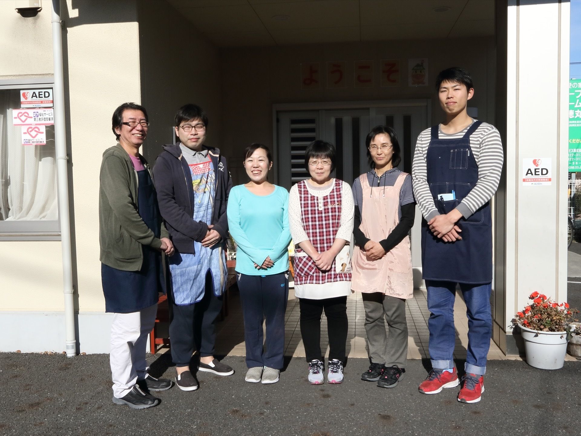 施設玄関前で、年齢に幅のある男女のスタッフが、作業着スタイルで両手を前で組み、こちらに微笑みかけている。