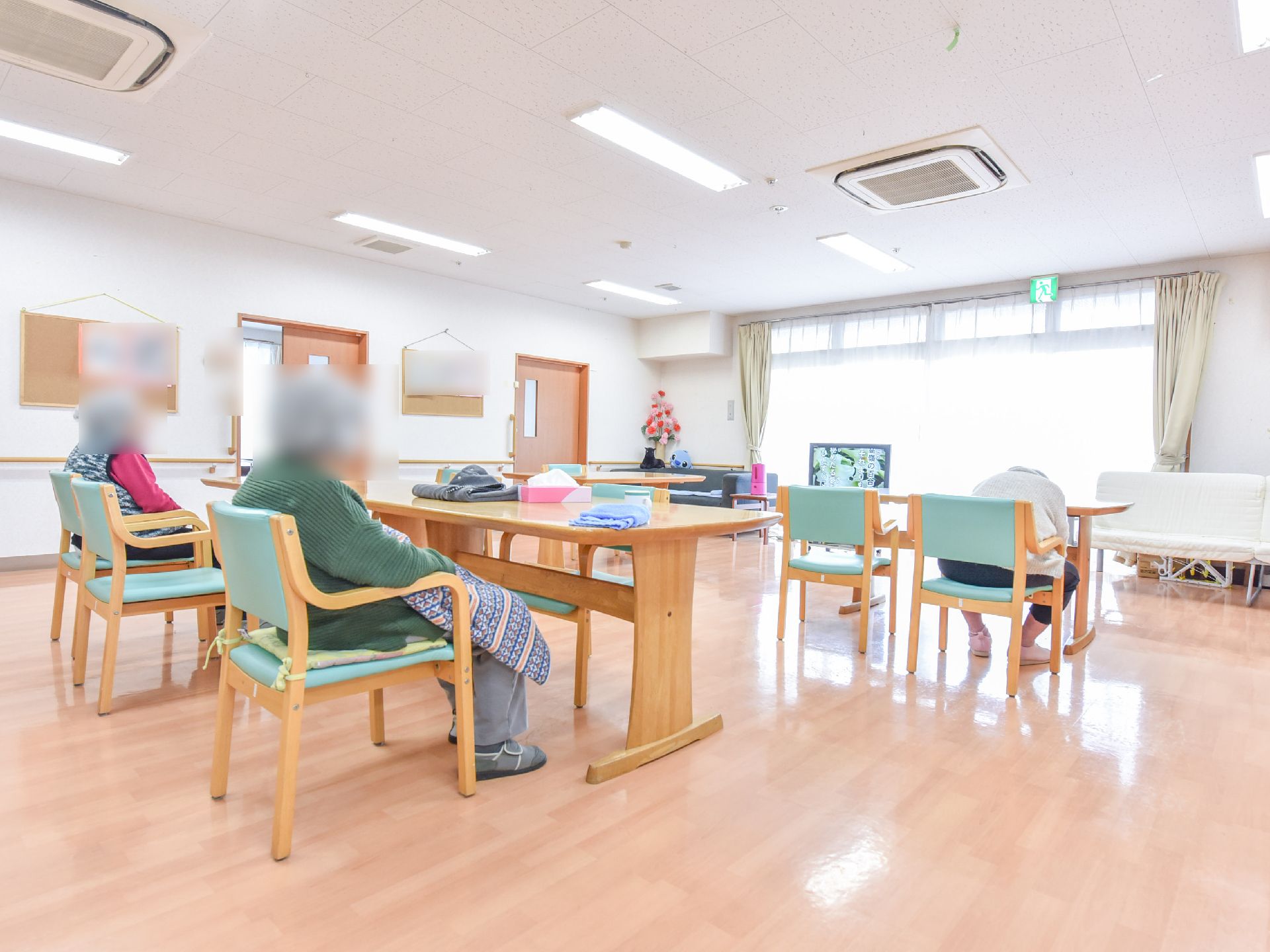 リビングは白壁にフローリングの床のホールになっている。木目調のデザインのテーブルが4つと椅子が8つ置かれ、入居者が座っている。