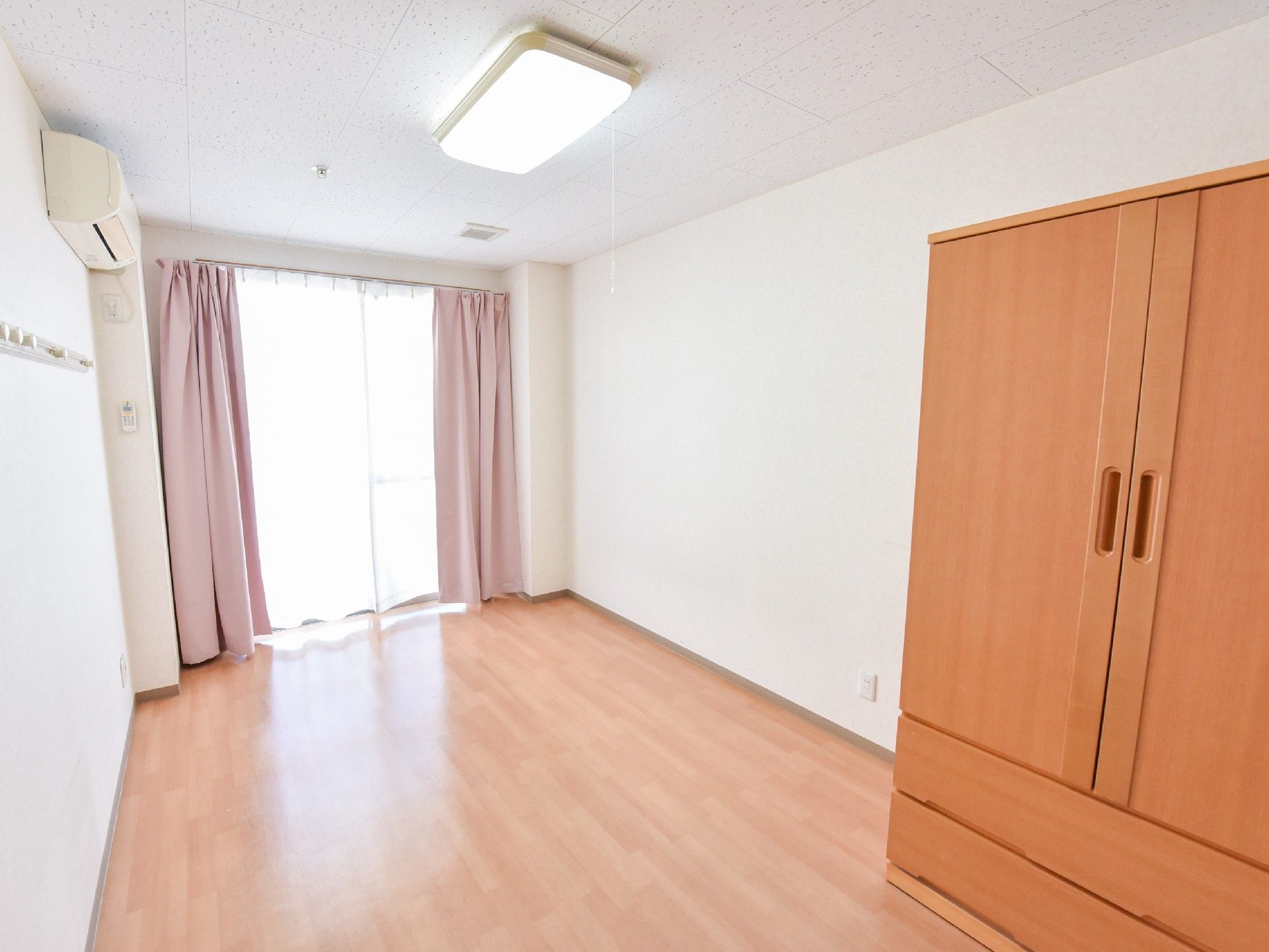 居室は白壁と茶色のフローリング。奥には窓がありカーテンが設置されている。エアコンとクローゼットも完備されている。