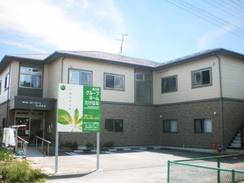 ベージュと茶色の外壁の二階建て建物で、玄関前には手すり付きの階段とスロープがある。建物前に駐車スペースがある。