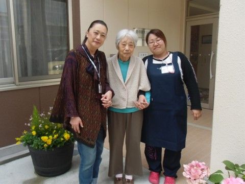 施設建物前で、紺色のエプロンに名札を付けた女性スタッフと、来訪者様に両脇を支えられて、入居者様がこちらを向いて立っている。