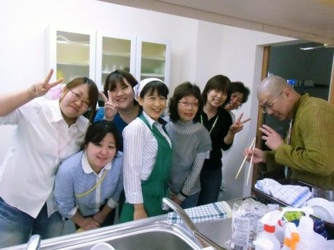 対面型キッチンのシンクの前で、緑のエプロンに三角巾を身につけたスタッフを囲んで、入居者様と名札をつけたスタッフが笑顔で並んでいる。