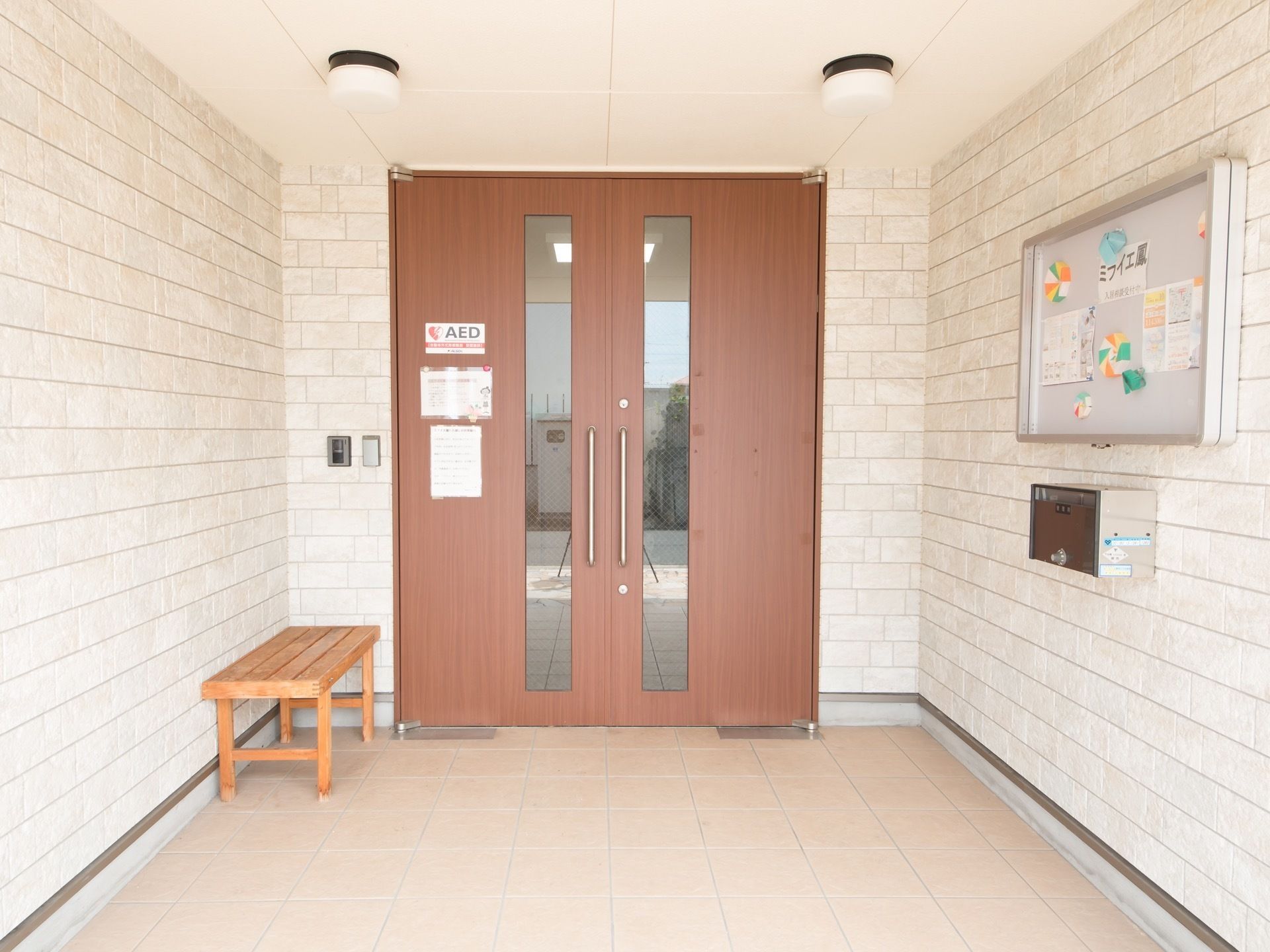 両開きの玄関扉である。ベンチが置かれ、反対の壁には掲示板が備わっている。また目立つ場所にAEDのステッカーが貼られている。