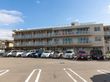 ３階建てのモダンな建物である。玄関前には広い駐車スペースがとられており、屋上には白いフェンスが見えている。