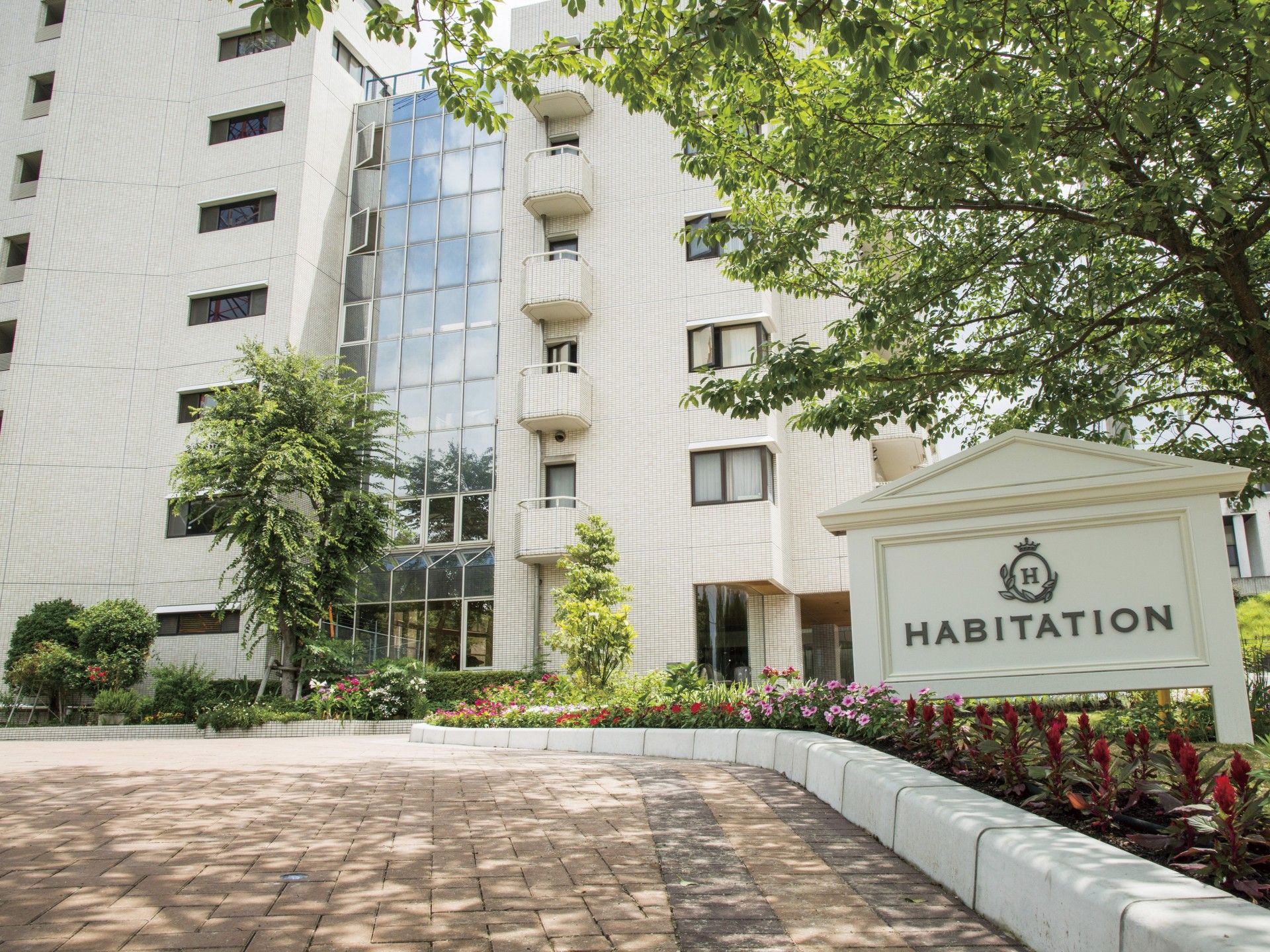 ６階建てのマンションとその後ろに８階建ての別の棟が建てられている。１階の施設前には駐車スペースがある。