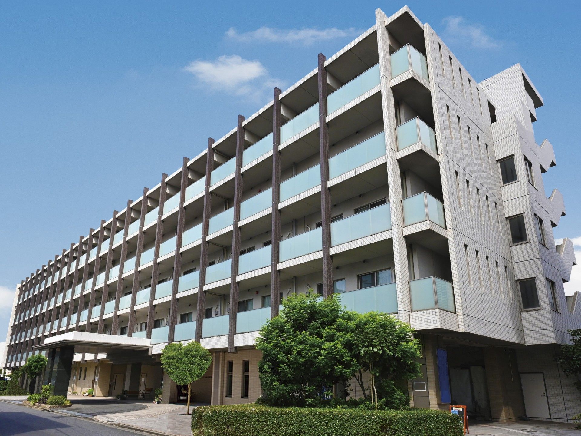 白壁の施設の外観。各居室に大きな窓とベランダが付いているのが見られる。玄関は奥まったところにあり、木の植え込みが目の保養になる。