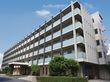 白壁の施設の外観。各居室に大きな窓とベランダが付いているのが見られる。玄関は奥まったところにあり、木の植え込みが目の保養になる。