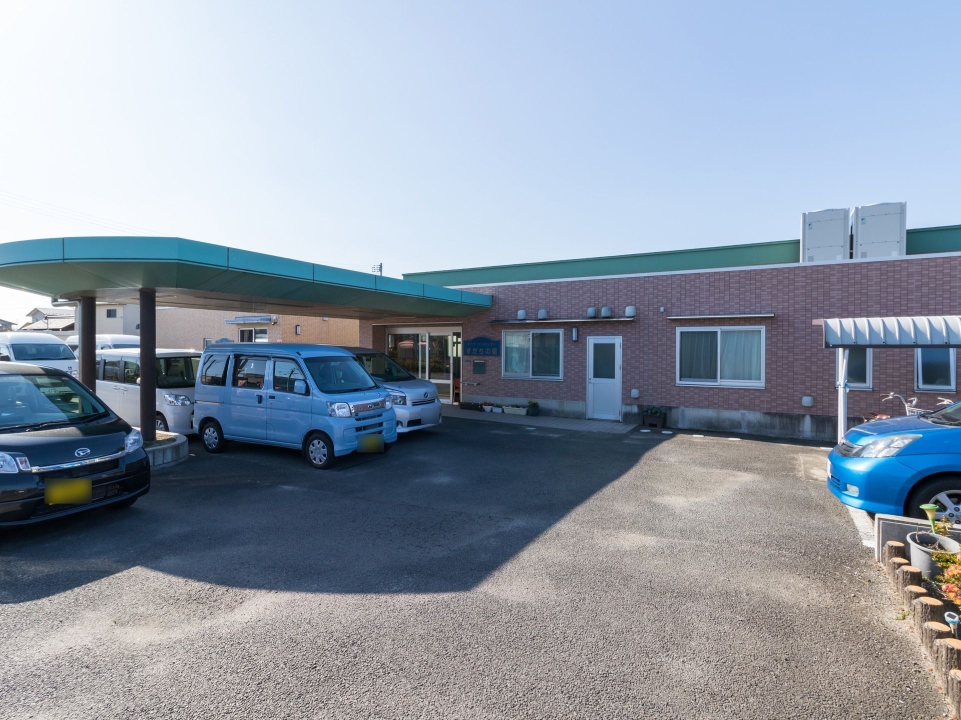 緑色の屋根と茶系のタイル調の外壁の平屋建ての建物である。敷地内には、車を停めるスペースがある。玄関ドア横には、表札がある。