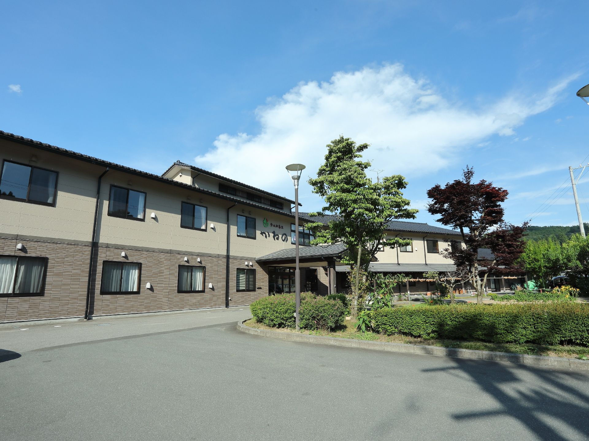 施設の建物は2階建てで、外壁の色はベージュ系である。建物の前面にある植え込みには複数の植物や木々が茂っている。