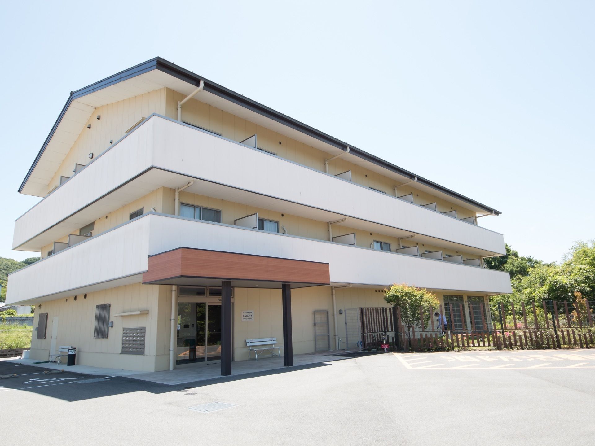 白地の壁に三角の屋根が特徴的な、コンパクトな大きさの建物である。入居者様の居室などが続く２階以上の階層には一続きのベランダが備わっている。