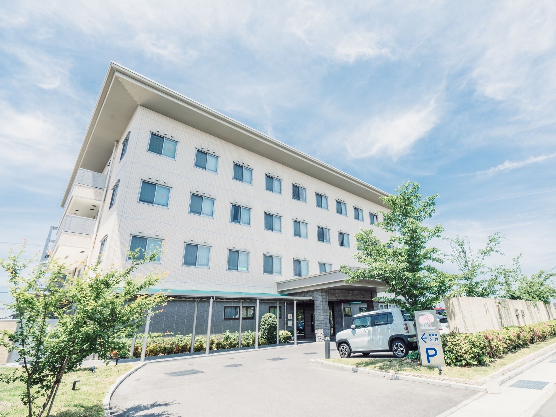 白色を基調とした４階建ての施設である。道路からの導入路に、駐車場入口と書かれた看板が設置されている。