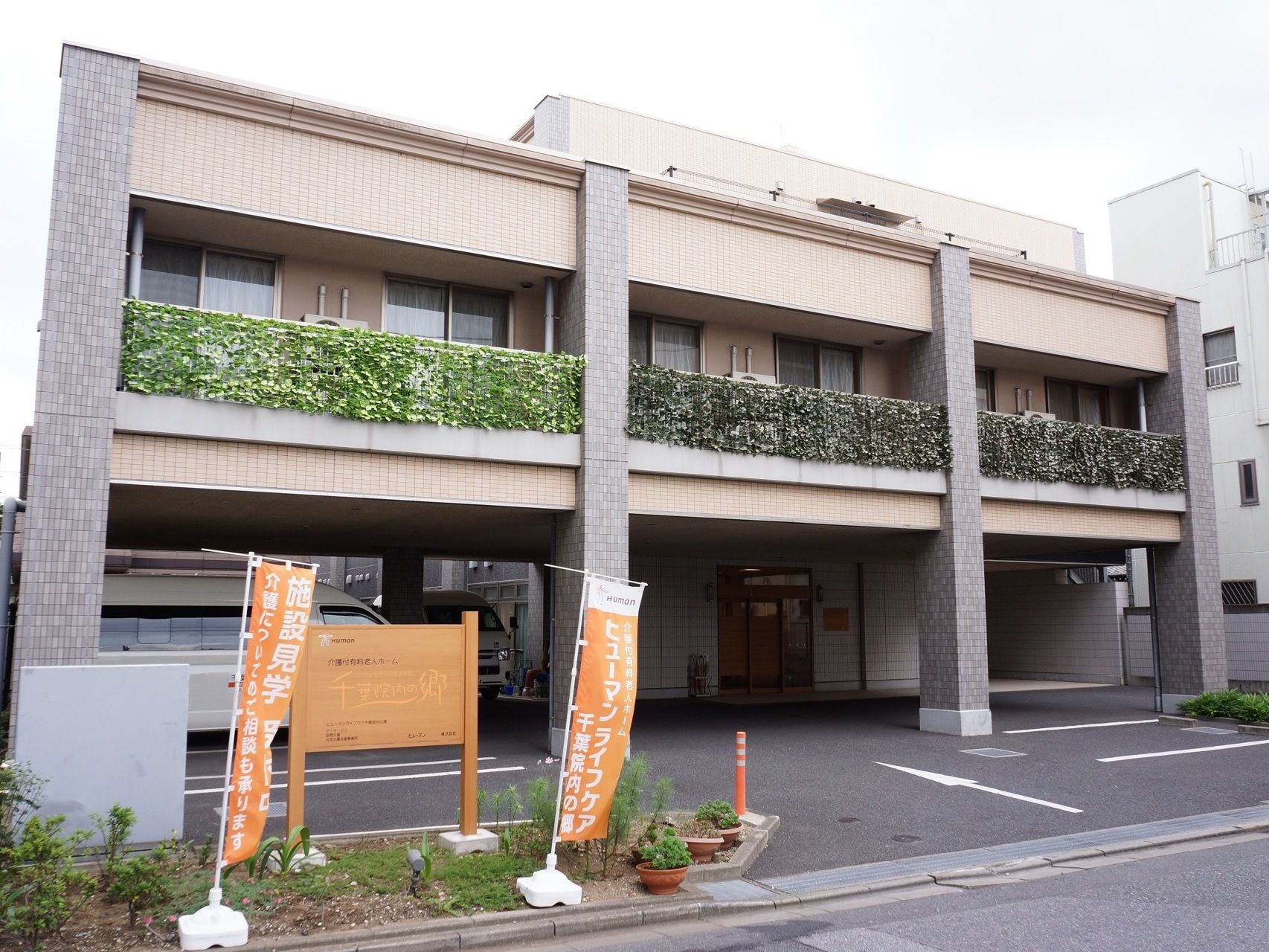 施設の入口には、施設名の書かれたのぼりが立っている。２階建ての施設となっており、べランダには緑がたくさん生い茂っている。