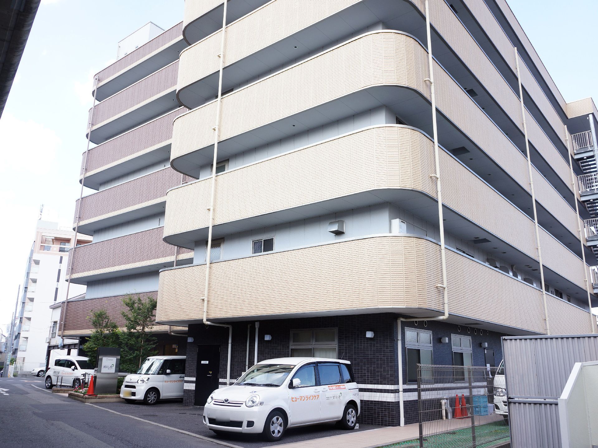 施設の一階部分に駐車場があり、車が数台停めてある。玄関の周りは、フェンスがあり、木が植栽してある。ベランダがついた建物である。