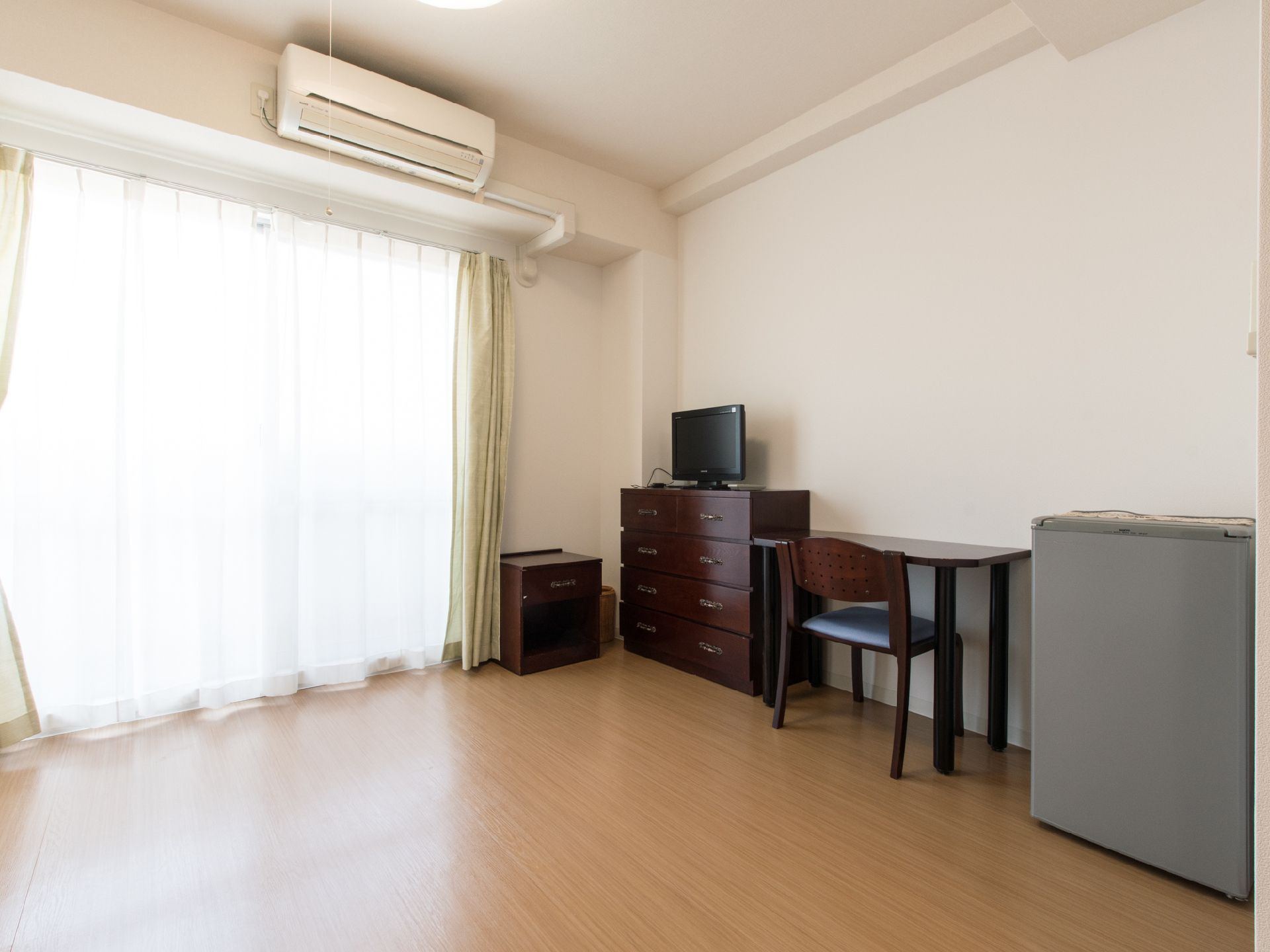エアコンのあるフローリングの部屋で、ベランダ窓がついていて陽光が差し込んでいる。壁際にチェスト、机、いすなどが置いてある。